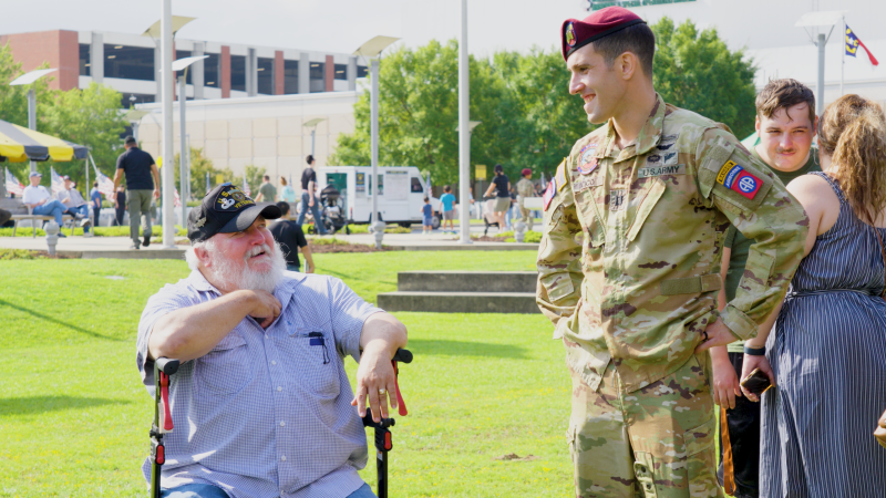 U.S. Army service member speaking with an older guest using a mobility chair during an outdoor community celebration in Fayetteville, NC.