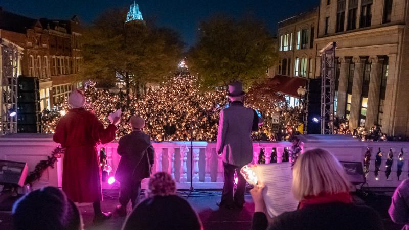 A crowd gathers in downtown Fayetteville under glowing lights as carolers in Victorian attire lead a holiday performance, filling the night with warmth and community spirit.