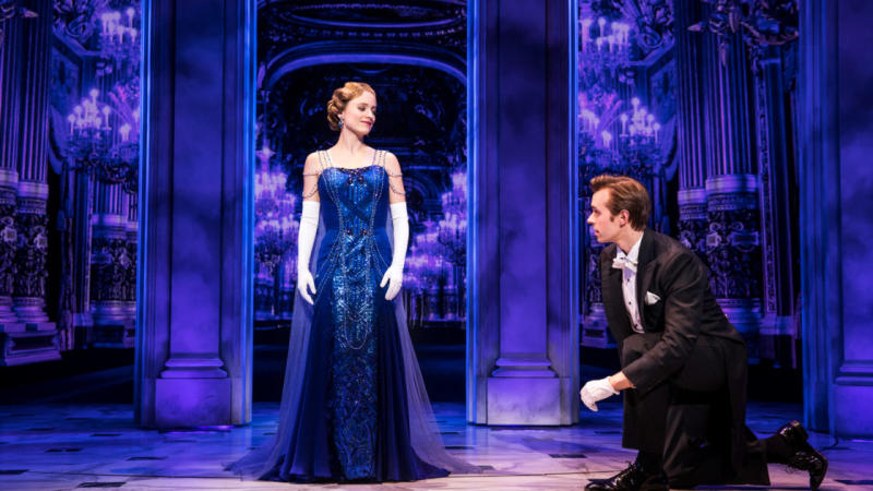 A woman stands on stage in a blue gown as a man kneels next to her in a performance of Broadway's Anastasia