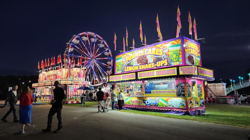 Image shows food booths and carnival rides lit up against a dark night sky at the Morgan County Fall Foliage Festival, held at the Morgan County Fairgrounds.