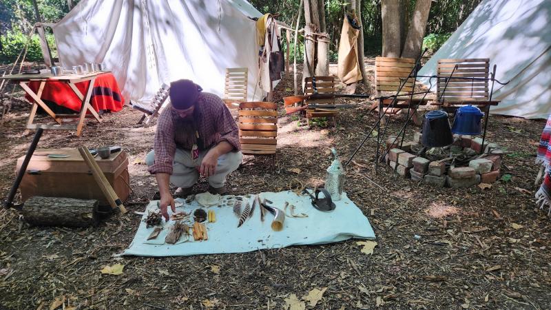A historic reenactor depicting the hunter/trapper era of American frontier history talks about his lifestyle and shows off various tools displayed on the ground during the Old Town Waverly Park Festival in Morgan County, Indiana.