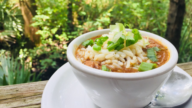 A white bowl filled with gumbo sits on a white plate on an outdoor table in front of green leaves and plants from Palmettos in Slidell