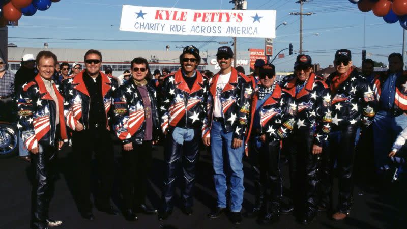 participants of the Kyle Petty Charity Ride pose for a photo under a banner