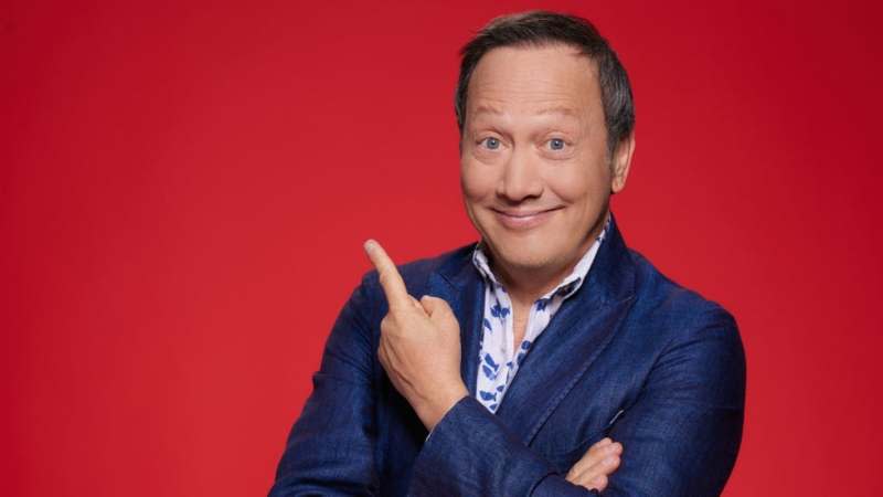 Rob Schneider is photographed smiling against a red background