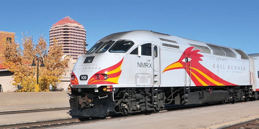 The New Mexico Rail Runner train stands still at the Alvarado Transportation Center, with the Downtown Albuquerque skyline in the background.