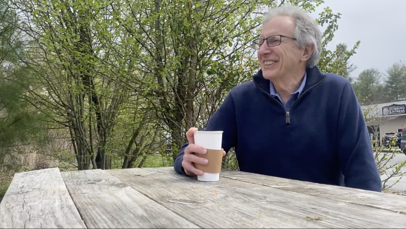 Dr. Ray Russell sits at a wooden picnic table with a coffee in one hand.