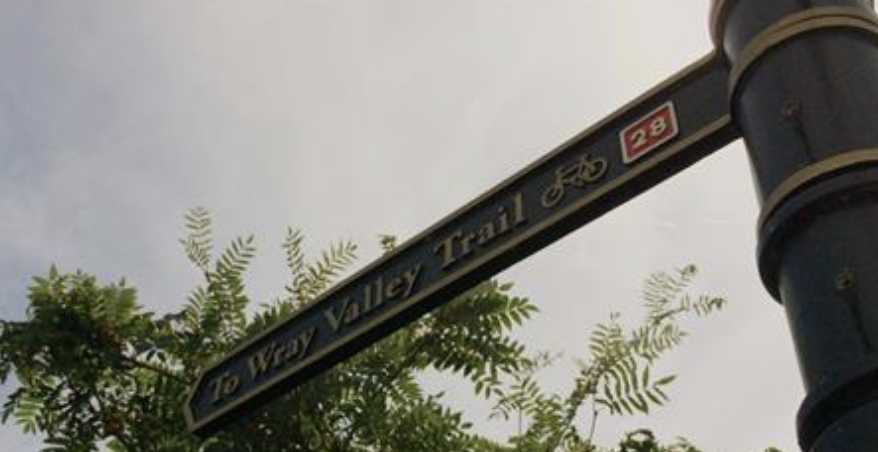 Signpost reading "To Wray Valley Trail" with a bicycle icon and number 28, set against a bright sky and lush green leaves, suggesting a scenic bike route.