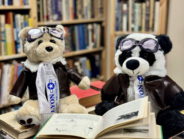 Adorable Lyon Air Museum plush toys, a panda and a bear dressed as aviators with leather jackets, scarves, and goggles, sitting among open aviation books in a cozy library setting