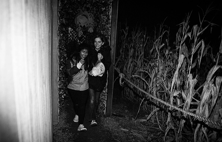 Two women walk through a dark corn maze, holding onto each other for support. Tall corn stalks surround them, creating a spooky atmosphere.