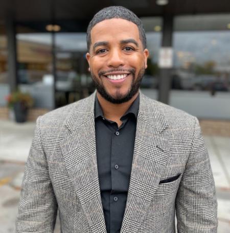 Man stands and smiles for a photo. He's wearinga grey suit with a black button up shirt underneath.