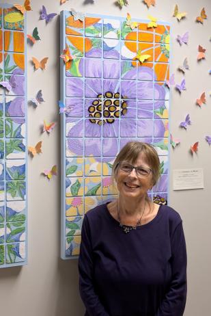 Local artist Sandra Cooper poses with the community mural that she designed, displayed at Canopy Cancer Survivorship Center at Hermann Memorial - The Woodlands. Cooper is wearing a dark long-sleeved shirt, a floral bead necklace, and purple classes. The mural consists of tiles painted with brightly colored flowers, surrounded by paper butterflies with hopeful messages on their wings.