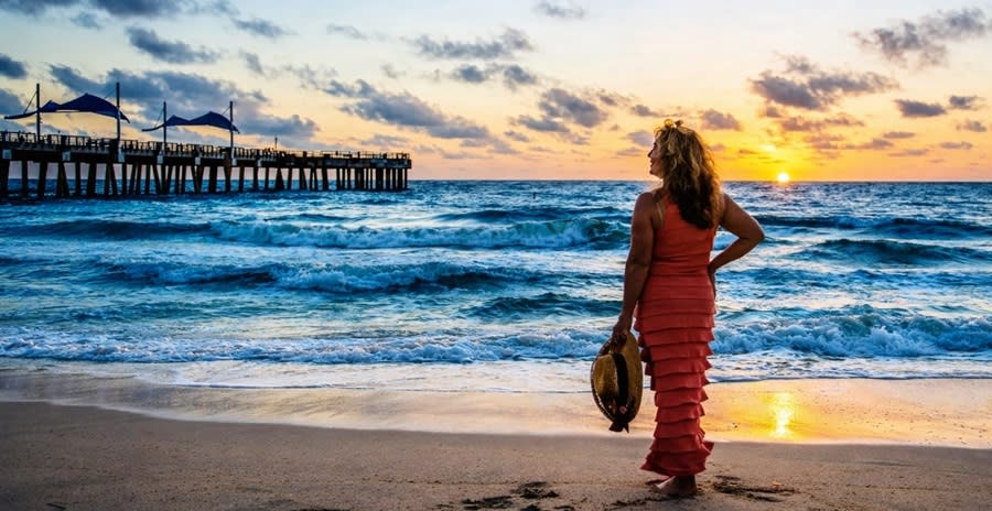 A woman standing at the ocean's edge watching the sunrise over the Pompano Beach Pier