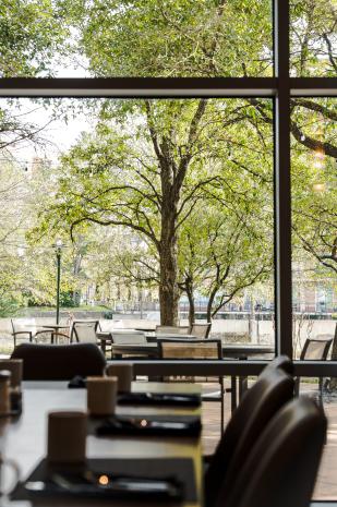 The camera overlooks the Acqua Patio through the Acqua Restaurant window. Visible from this angle are three round patio tables, each set with 2-4 comfortable patio chairs. The view also includes a lamp post, glimpses of The Waterway, and a number of healthy green trees wrapped in Christmas lights (off, given the daylight).