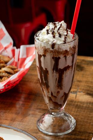 A clear milkshake glass is blended full of a chocolate shake with chocolate syrup and whipped cream. In the background is a basket of fried jalapeno slices.