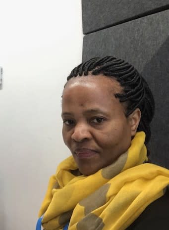 Close-up of a person with braided hair sitting indoors near acoustic wall panels, wearing a yellow hoodie, with part of a chair and a white wall visible in the background.