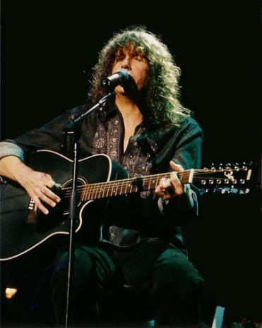 A close-up of Randy Jackson performing. He has long, curly brown hair, is wearing a dark denim shirt with brown embroidery down the chest and the sleeves rolled up to his elbows. He is strumming a black acoustic guitar and singing into a microphone. He is sitting on a stool, presumably on stage, though the background is entirely black.