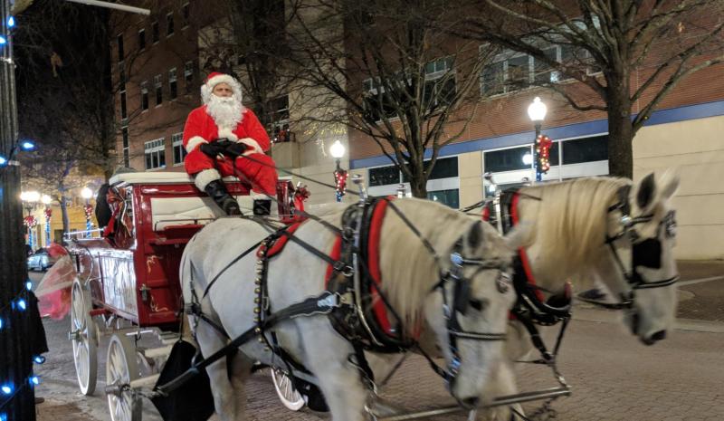 Santa Claus sits atop a red horse-drawn carriage on a brick-lined street in downtown Fayetteville, surrounded by twinkling lights and festive décor.