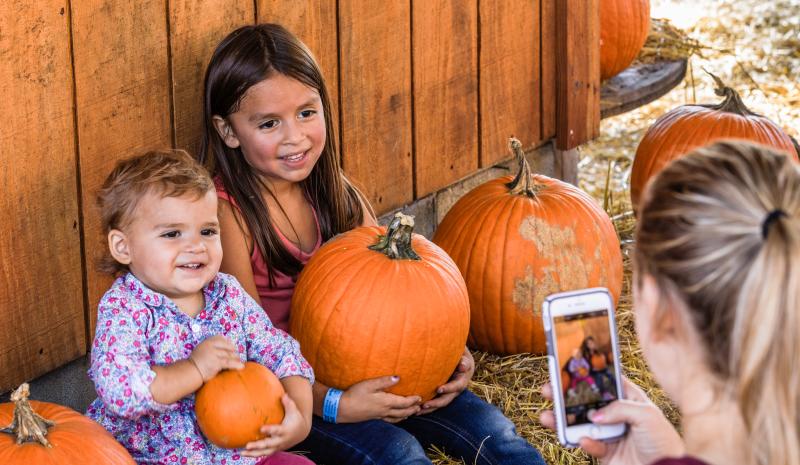 Two young children sit among pumpkins smiling while an adult takes their photo on a smartphone, capturing a festive fall selfie moment in Fayetteville.