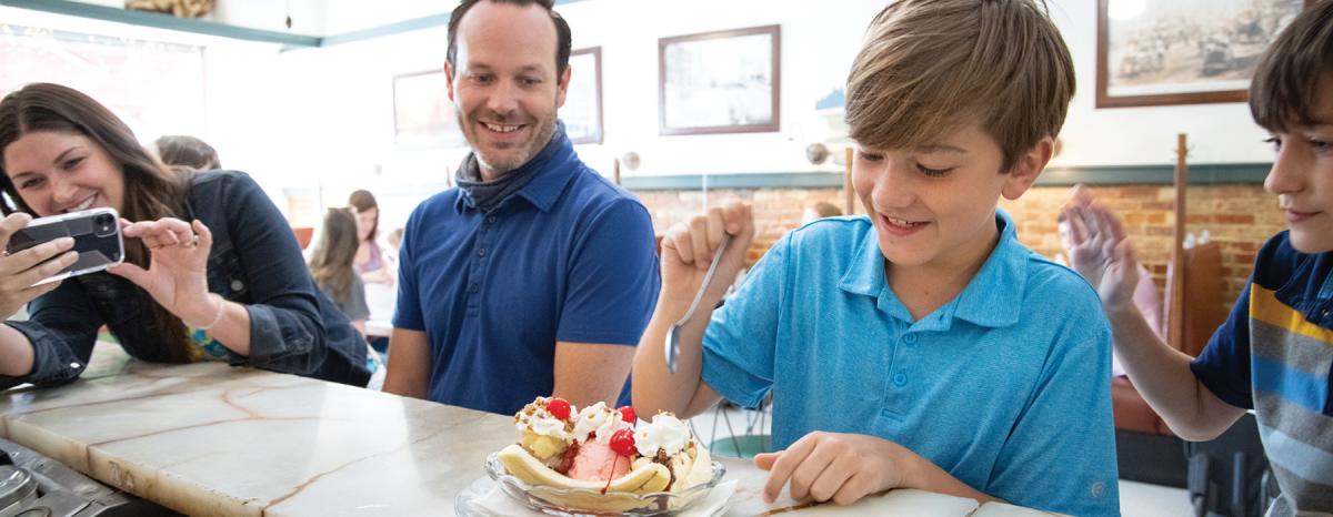 A mother takes a photo of her son sitting between his dad and brother while he digs into a banana split at Old Mill Tasty Shop in downtown Wichita