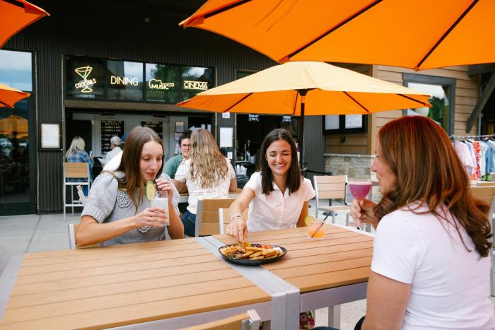 Three women enjoy drinks and snacks at a wooden table under bright orange umbrellas. A lively outdoor dining scene is visible in the background.