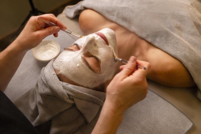 A person receiving a facial treatment, with a white mask applied, while a therapist uses brushes. Soft towels and a bowl of cream are visible.