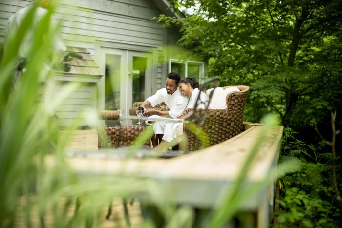 A couple toasts with wine glasses in white robes on a wicker couch outdoors.