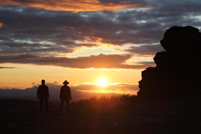 Two men watching sunset on the horizon with boulders in foreground in
