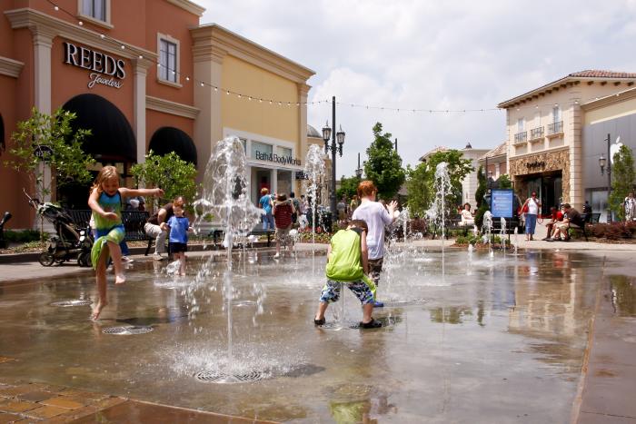 Children play in the interactive splash pad at Bridge Street Town Centre in Huntsville, running through fountains and water jets on a sunny day. Shops, outdoor seating, and landscaped walkways surround the plaza as families enjoy the outdoor shopping and entertainment district.