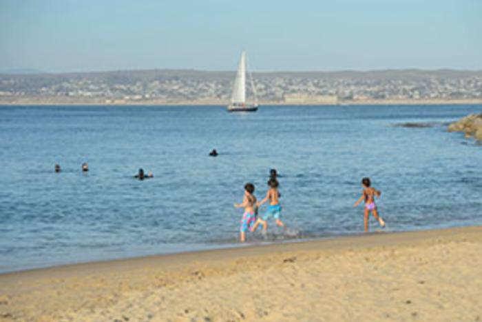 Children run into the ocean with sailboat in the background in Monterey County, CA