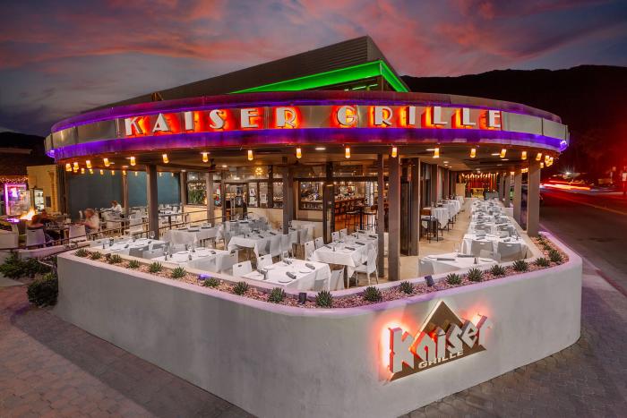 An exterior view of Kaiser Grille restaurant at dusk, showcasing neon signage, outdoor seating with white tablecloths, and a vibrant sunset sky.