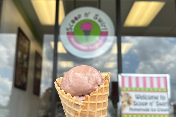 A hand holding a chocolate ice cream cone in front of an ice cream shop
