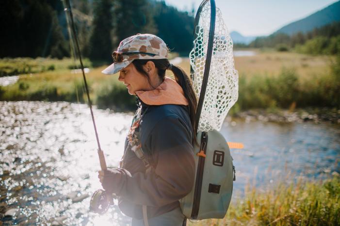 A person stands by a shimmering river, holding a fishing rod, with a net on their back and a scenic landscape of trees and mountains behind.