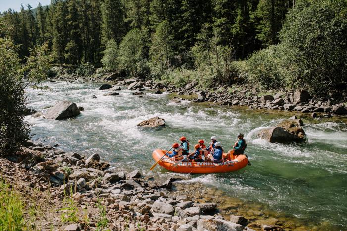 A bright orange raft with six people navigates a rushing river, surrounded by lush green trees and rocky banks. The scene captures an adventurous outdoor experience.