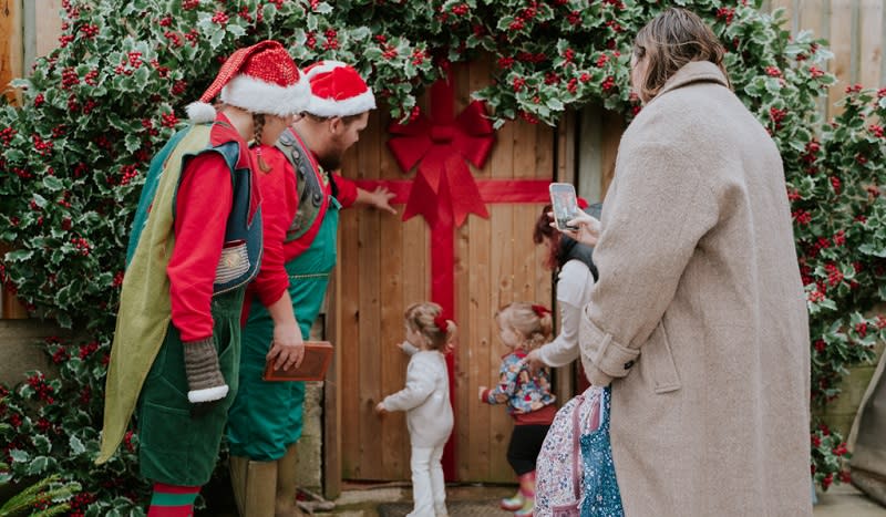 Two little children push open the door to Santa's grotto, watched by two elves and their parents