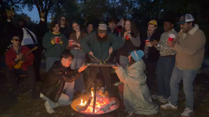 A group of people gather outdoors around a campfire at dusk, many holding red cups and watching. In the center, two people kneel on either side of a small tabletop instrument while another person stands behind it, playing it with mallets. A person off to the side plays a small guitar or ukulele, adding to the casual, festive atmosphere.