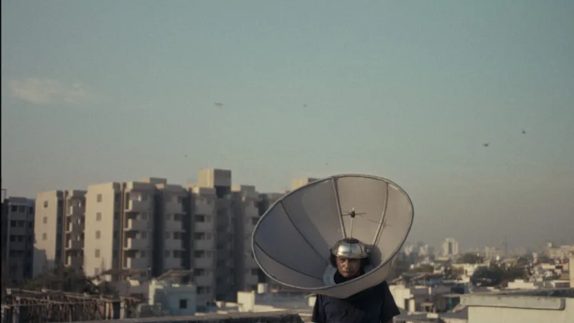 A person stands on a rooftop in an urban area, wearing a large satellite dish around their head like a helmet. In the background, there are mid-rise apartment buildings and a hazy city skyline under a pale sky.