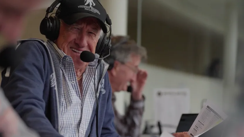 Bob Uecker wearing a baseball cap and headset microphone smiles while seated at a table, holding a sheet of paper. Another person sits slightly behind him, out of focus, appearing to read or concentrate. The setting looks like a broadcast or commentary area.