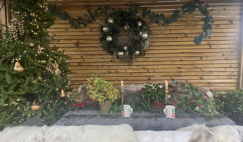 A firelit wooden shelter with fur blankets on the seats, decorated with Christmas tree and wreath