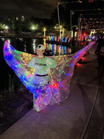 A woman, 30s, cheers on IRONMAN athletes. She is wearing a T-shirt tie-dyed white and neon-green, dark athletic pants, sneakers, and a white ballcap. In her hands are two wands waving a swath of metallic fabric (like butterfly wings) lined with multi-colored LED lights. The sky is dark, and behind her is the "Hippie Hollow" no-walking zone, lit up and crowded with other fans hyping up the runners as they approach the finish line.