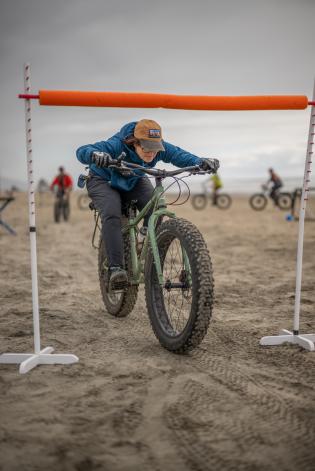 A person goes under the limbo bar on their fat tire bike on the beach.