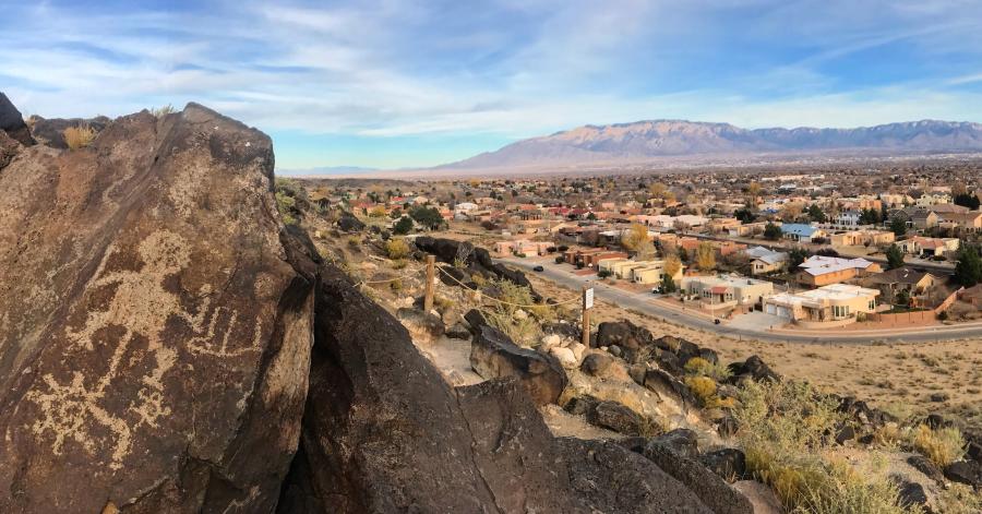 In the foreground is a rock with carved petroglyphs at the Petroglyph National Monument. The city of Albuquerque and the Sandias are in the distance.