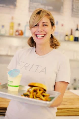 Chef Eve Aronoff Fernandez stands in her restaurant Frita Batidos and holds a tray containing a lemonade with a blue umbrella and a frita burger covered in shoestring french fries.