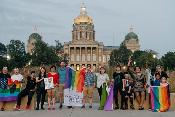 Group of people holding rainbow flags and signs in front of the Iowa State Capitol