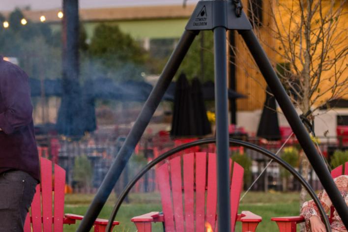 People sitting around a fire in red Adirondack chairs, in the back there is a building and an outdoor stage