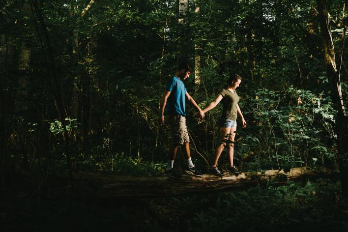 A young man and young woman walking on a log, holding hands in the forest