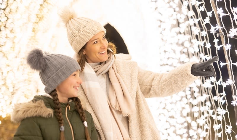 A mother and daughter wearing wooly hats, scarves and gloves in a tunnel of fairy lights