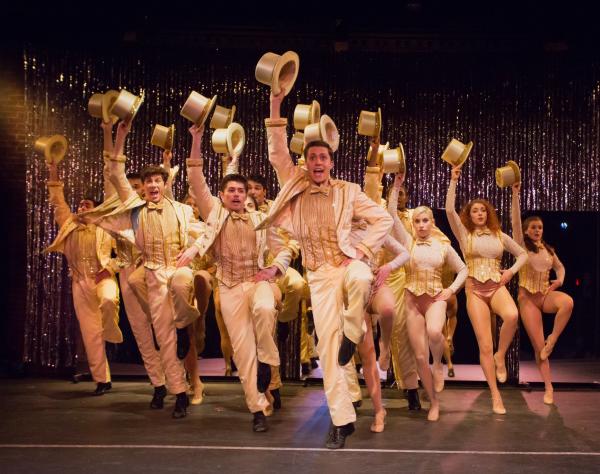 A group of men and women perform a scene from a play at the Clear Space Theatre in Rehoboth, Delaware. The actors are dressed in gold tuxedos while the actresses are in gold vests. All of them are holding gold top hats while dancing and singing.