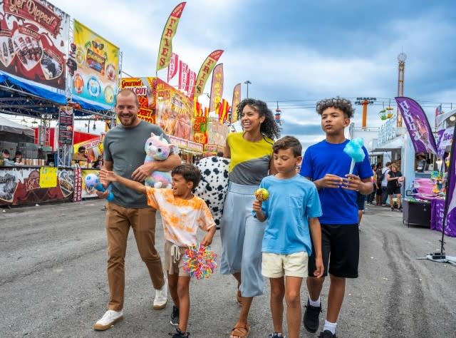 A smiling family with three kids walks happily at the Great New York State Fair, holding cotton candy, toys, and prizes. Bright vendor stalls and banners line the background.