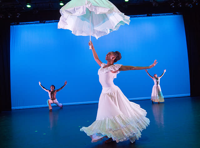 Two female and one male dancer dressed in white dance in front of a blue backdrop at the Alvin Ailey American Dance Theater