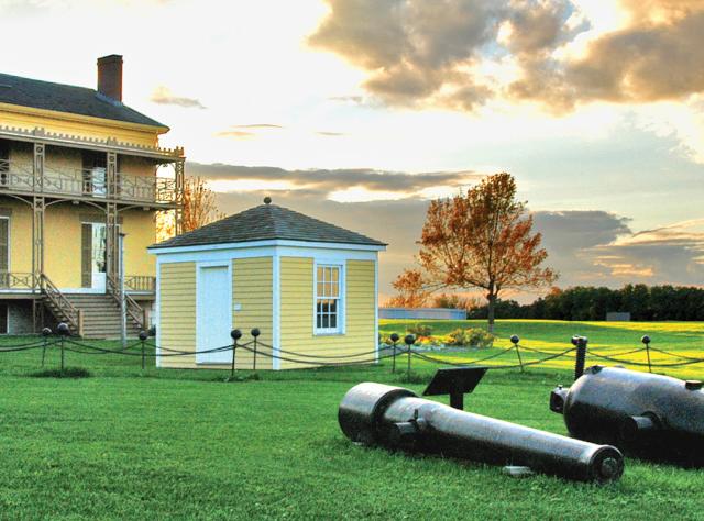 Two cannons resting on the field at Sackets Harbor Historic Site with a house and out-house in the background
